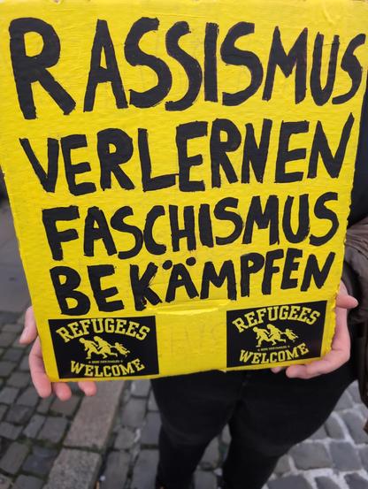schwarze Schrift auf gelbem Plakat:

Rassismus verlernen
Faschismus bekämpfen

Darunter 2 Aufkleber: "Refugees welcome".
