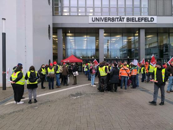 Ein Haufen Menschen vor dem Haupteingang der Universität Bielefeld. Viele tragen gelbe Warnwesten und rote Mützen der Gewerkschaft ver.di.

(aktuelles Foto vom 14. Januar 2026 um 10:28 Uhr)