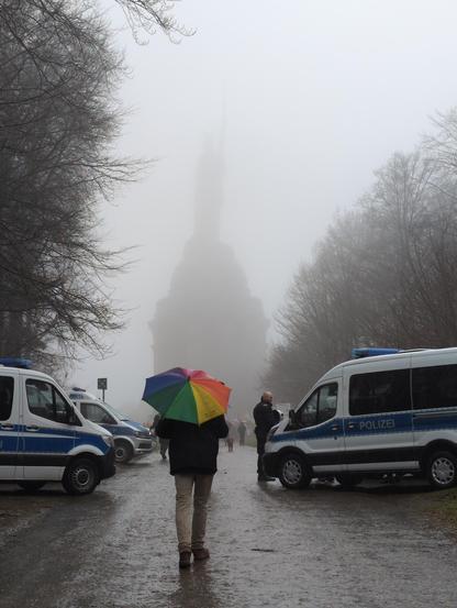 Ein Gegendemonstrant mit buntem Regenschirm auf dem Weg zu Hermannsdenkmal, das wegen des Regenwetters nur schemenhaft zu erkennen ist. Links und rechts Polizeiwannen.