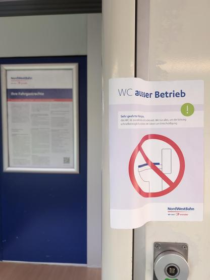 Die Tür zur einzigen Toilette der RB 85 (74937) der NordWestBahn ist mit einem Hinweis zugeklebt, auf dem steht: "WC außer Betrieb". Im Hintergrund ein Plakat, das über Fahrgastrechte aufzuklären vorgibt.