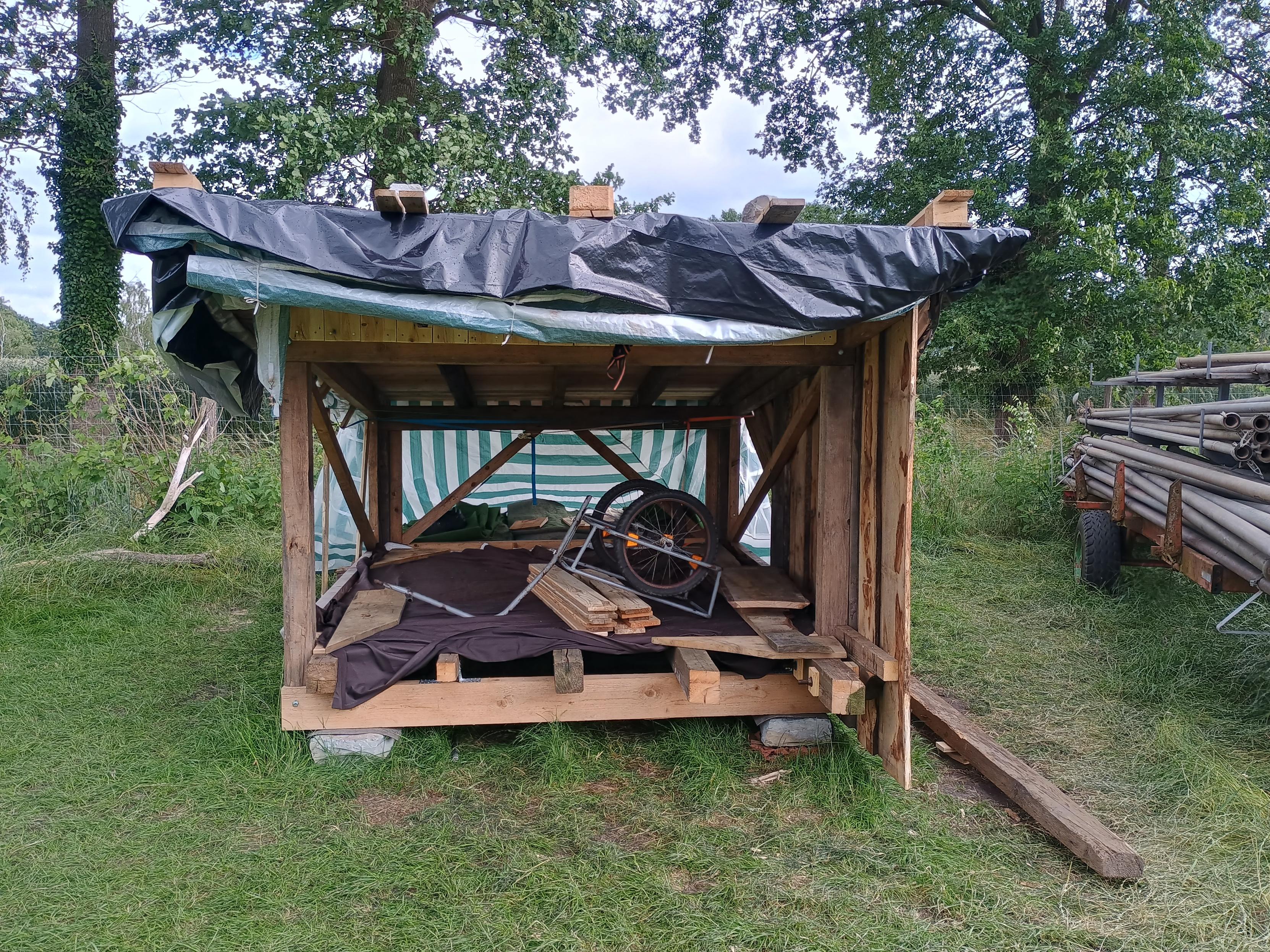 Ein Foto einer Shelter-Hütten-Baustelle auf einer Wiese. Die halbfertige Holzhütte ist temporär mit verschiedenen Planen abgedeckt. Im inneren liegt brauner Staooff, daraf Bretter und ein Fahrradanhänger.