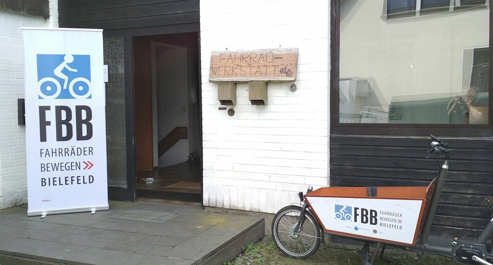 Ein Foto des Eingangs der FBB Werkstatt in Brackwede. Zu sehen ist ein Banner mit dem Logo von FBB "Fahrräder bewegen Bielefeld und ein Lastenrad, ebenfalls mit dem Logo von FBB.