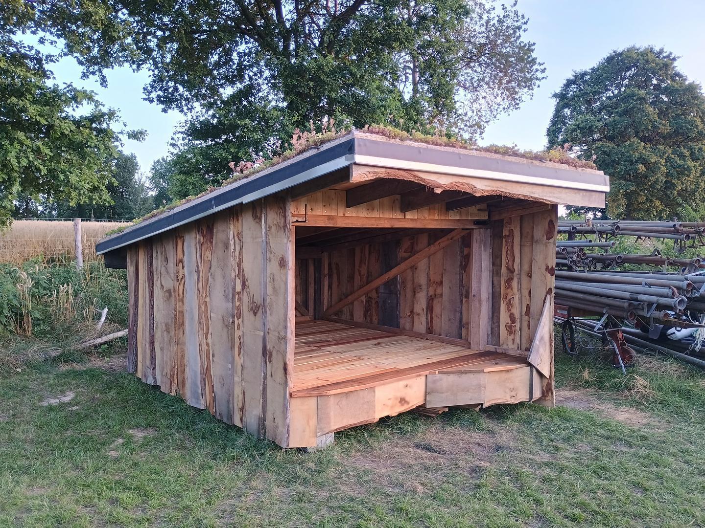 Ein Foto von einer rustikalen Shelter-Hüttte mit Gründach, auf einer Wiese. Im Hintergrund Bäume. Rechts daneben ein landwirtschaftlicher Rohr-Anhänger zur Bewässerung.