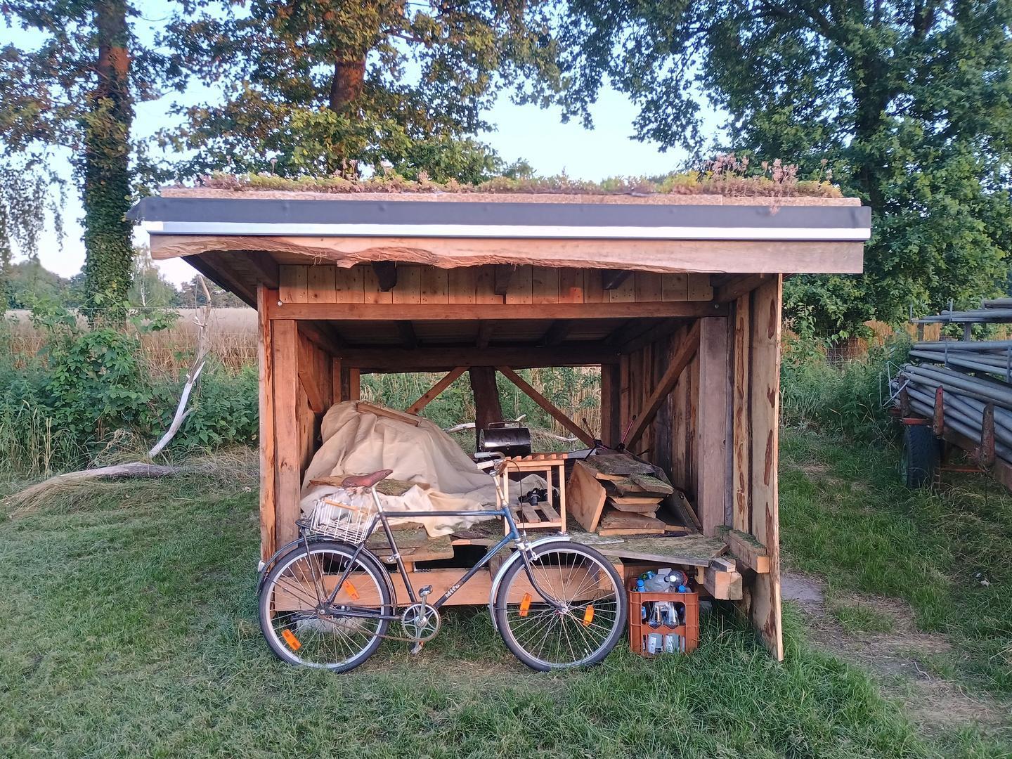 A foto of a shelter-hut with a green plant roof on a meadow. Trees in the background. Inside the shelter you see stuffed materials and the rear wall is still needs to be closed. A bike and some water bottles are at the front edge of this cosy oakwood building for hiking and biketravels.