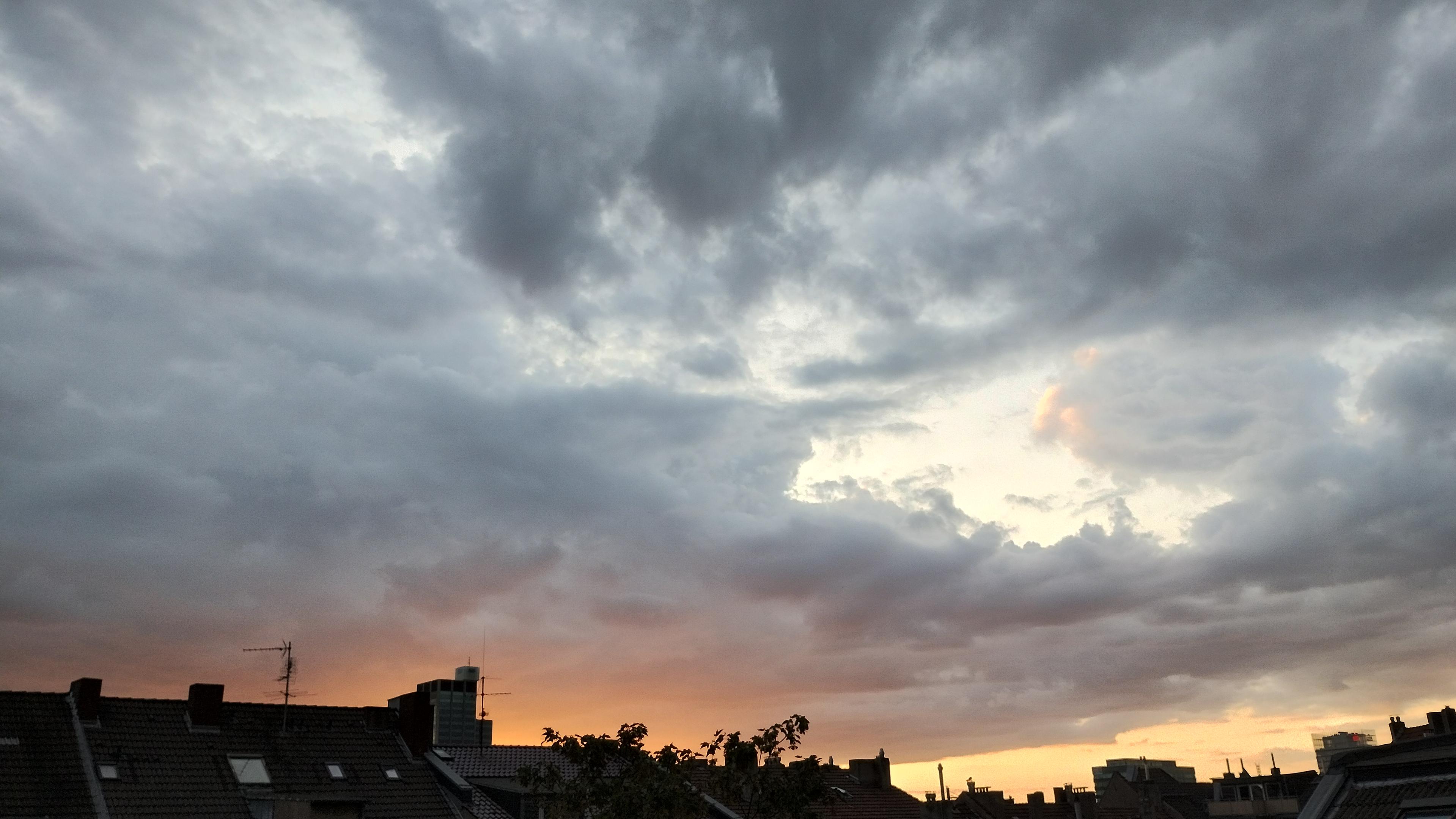Über den Dächern der Mietshäuser ist der grau bewölkte Abendhimmel. Die Wolkendecke ist nicht ganz geschlossen. Vom Sonnenuntergang sind nur noch Reste in orange und gelb zu sehen.