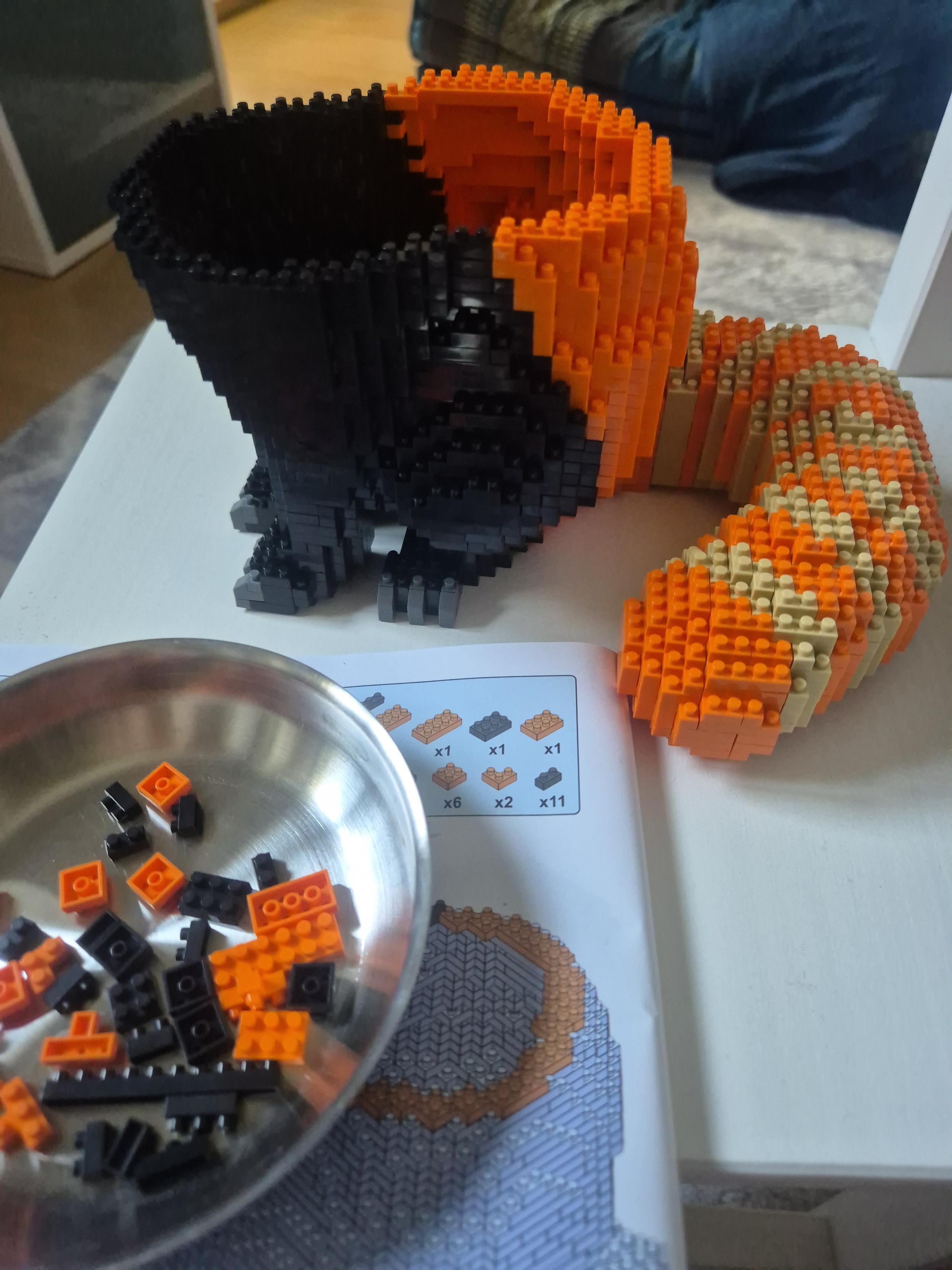 Auf einem kleinen weißen Stuhl steht ein halb fertiges Tier aus schwarzen, orangenen, beigen und grauen sehr kleinen Klemmbausteinen. Man sieht drei Pfoten eines sitzenden Tieres mit buschigem Schwanz. Etwa die Hälfte des Rumpfes sowie der Kopf fehlen noch.
Davor ist eine Anleitung sowie eine Schale mit einigen Klemmbausteinen.