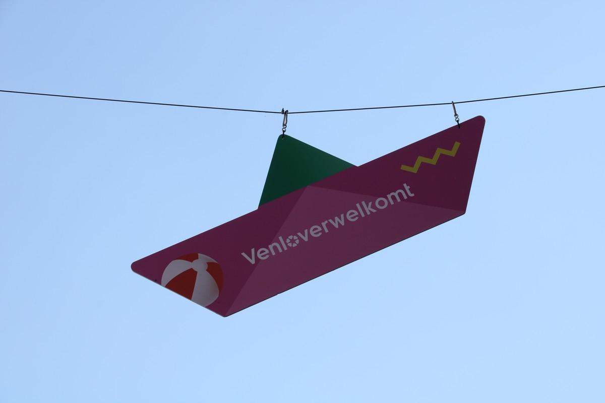 Foto im Querformat. Vor einem blauen HImmel hängt ein gefaltetes Schiffchen, aber aus Kunststoff statt aus Papier. Das „Segel“ ist grün und der „Schiffskörper“ rosa/pink. Auf dem Schiffskörper steht in weiß „Venloverwelkomt“. Links von der Schrift ist ein aufblasbarer Bade-Ball abgebildet, rechts ein gelbes Zick-Zack-Muster, das wohl Wasser darstellen soll.